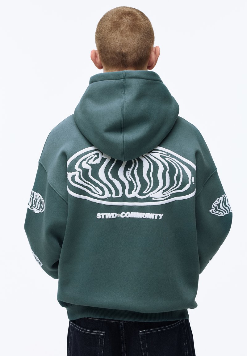 Personne portant un sweat à capuche bleu sarcelle avec un motif abstrait blanc audacieux et le texte « STWD COMMUNITY » au dos, tournée de dos à la caméra.