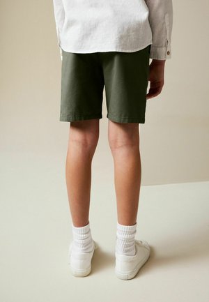 Rohelised puuvillased lühikesed püksid, sääre kohal, lihtsa disainiga. Kandmisel valgete tossude ja sokkidega, vaadatuna tagant neutraalse tausta ees.