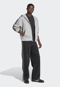 Šedá mikina na zips s kapucňou, so šnúrkami a čiernymi pruhmi na rukávoch, v kombinácii s čiernymi voľnými nohavicami so bočnými pruhmi a čiernymi topánkami.