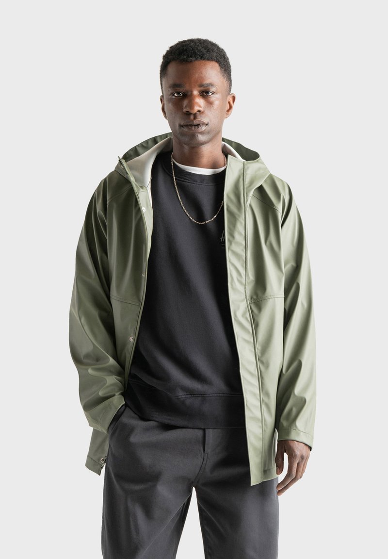 Vattentålig grön jacka med justerbar huva, mjukt innerfoder och sidofickor, som bärs över en svart sweatshirt och mörka byxor.