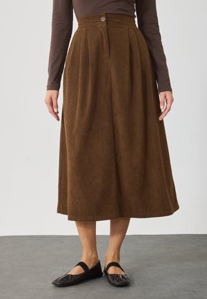 Jupe midi marron en velours côtelé taille haute avec fermeture à boutons, portée avec un haut à manches longues marron foncé et des ballerines Mary Jane noires.