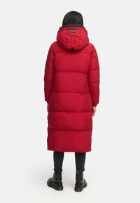 Rød puffjakke med hætte, der har et quilted design og forlænget snit. Jakken er parret med sorte leggings og snørebåndsstøvler.