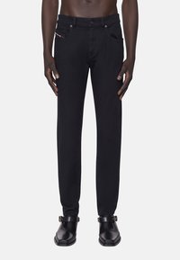 Man som bär slim-fit svarta jeans och svarta läder ankellång stövlar med spännen, står mot en neutral bakgrund.