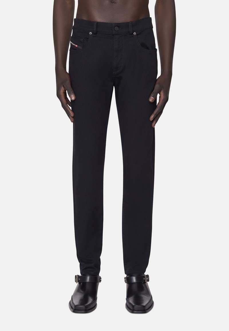 Man som bär slim-fit svarta jeans och svarta läder ankellång stövlar med spännen, står mot en neutral bakgrund.