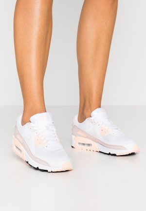 Jambes portant des baskets Nike Air Max blanches et rose clair avec une semelle visible à coussin d'air, debout sur un sol clair uni face à un fond clair.