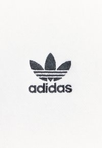 Vitt bomullstyg med en svart broderad Adidas-logotyp, med tre bladliknande detaljer och tre horisontella ränder under namnet.