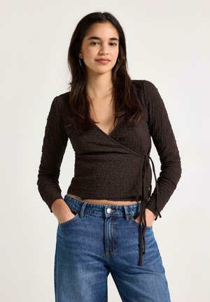 Kvinde med langt mørkt hår iført en tekstureret sort omslagsbluse med lange ærmer og blå jeans, stående foran en ensfarvet baggrund.