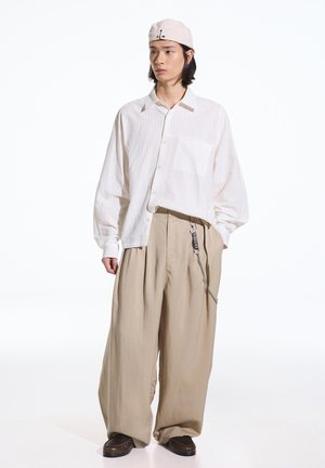 Jeune personne portant une chemise blanche oversize, un pantalon large beige avec un accessoire chaîne, des chaussures marron foncé et une casquette beige clair, debout devant un fond blanc.