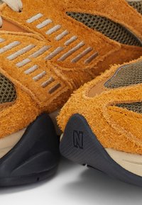 Gros plan sur des baskets texturées orange avec des panneaux en mesh gris et des semelles noires ornées d’un logo en relief "N".