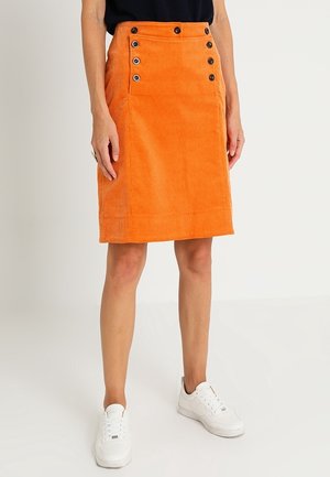 Kvinde iført en knælængde orange kordnedsat nederdel med sorte knapper foran, parret med hvide sneakers og en sort top.