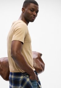 Camiseta ajustada beige, de mangas cortas. Jeans de mezclilla con una camisa de franela a cuadros atada a la cintura. Bolso de lona marrón.