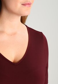 Primo piano del collo e della parte superiore del torace di una donna che indossa un top bordeaux a V su uno sfondo semplice.