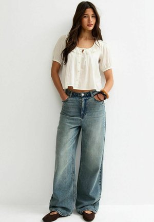 Woman stands against white wall wearing a white short-sleeve blouse, wide-leg faded blue jeans, brown flats, and bracelets on one wrist.