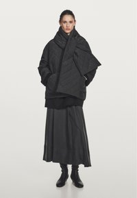 Black quilted jacket with a high collar, long sleeves, and front pockets, worn over a long black skirt and ankle boots.