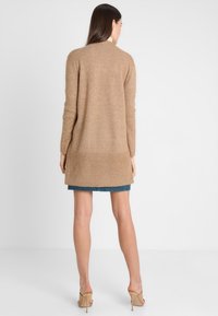 Cárdigan largo beige hecho de material de punto suave, con un dobladillo acanalado y un ajuste relajado, que se extiende por debajo de las caderas.