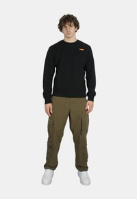 Felpa nera con polsini a coste, abbinata a pantaloni cargo verde oliva con più tasche e zip. Sneakers bianche completano l'outfit.