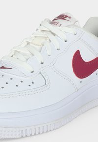 Λευκό αθλητικό παπούτσι Nike Air Force 1 με κόκκινο λογότυπο Nike swoosh, λευκά κορδόνια, διάτρητη μύτη και υφασμένο λευκό πέλμα.