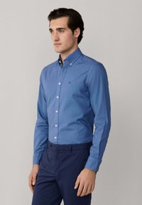 Junger Mann in einem blauen Hemd mit Knöpfen und dunkelblauer Hose steht vor einem einfarbigen Hintergrund und schaut leicht nach links.