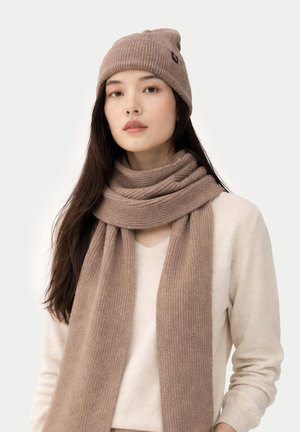 Young woman wearing a beige knit hat and matching long scarf over a cream sweater, facing forward with a neutral expression.