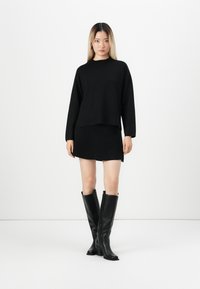 Maglione nero lavorato a maglia con maniche lunghe, sovrapposto a una mini gonna nera, abbinato a stivali alti in pelle nera, su uno sfondo neutro.