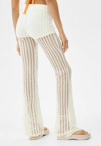 Białe dzianinowe spodnie w siateczkę o rozszerzającym się kształcie, z pionowymi paskami. Pasek w talii jest żebrowany, a materiał ma otwartą strukturę splotu.
