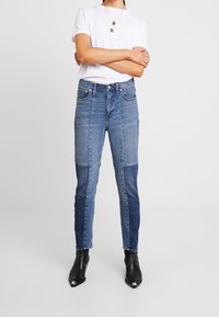 Jeans de denim azul claro con un diseño de patchwork que presenta diferentes tonos de azul. De cintura alta, ajustados, combinados con botas negras hasta el tobillo.