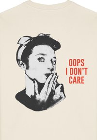Krēmkrāsas sporta krekls ar melnbaltu grafiku, kurā attēlots cilvēks ar pirkstu uz lūpām. Sarkanā krāsā teksts skan "OOPS I DON'T CARE."