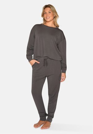 Blondynka stoi boso, uśmiechając się, ubrana w ciemnoszary top z długim rękawem i pasujące spodnie ze ściągaczem na jednolitym tle.
