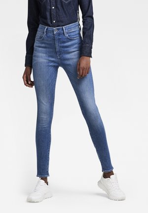 Personne portant un jean skinny bleu clair, une chemise en denim foncé et des baskets blanches, debout les jambes écartées devant un fond uni.