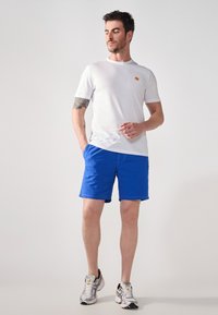 Biela tričko s krátkym rukávom s malým oranžovým logom, doplnené modrými šortkami a striebornými športovými topánkami. Hladká látka; voľný strih.
