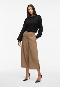 Svart strukturstickad tröja, beige vida byxor med sidofickor och svarta högklackade skor, presenterade mot en enkel vit bakgrund.