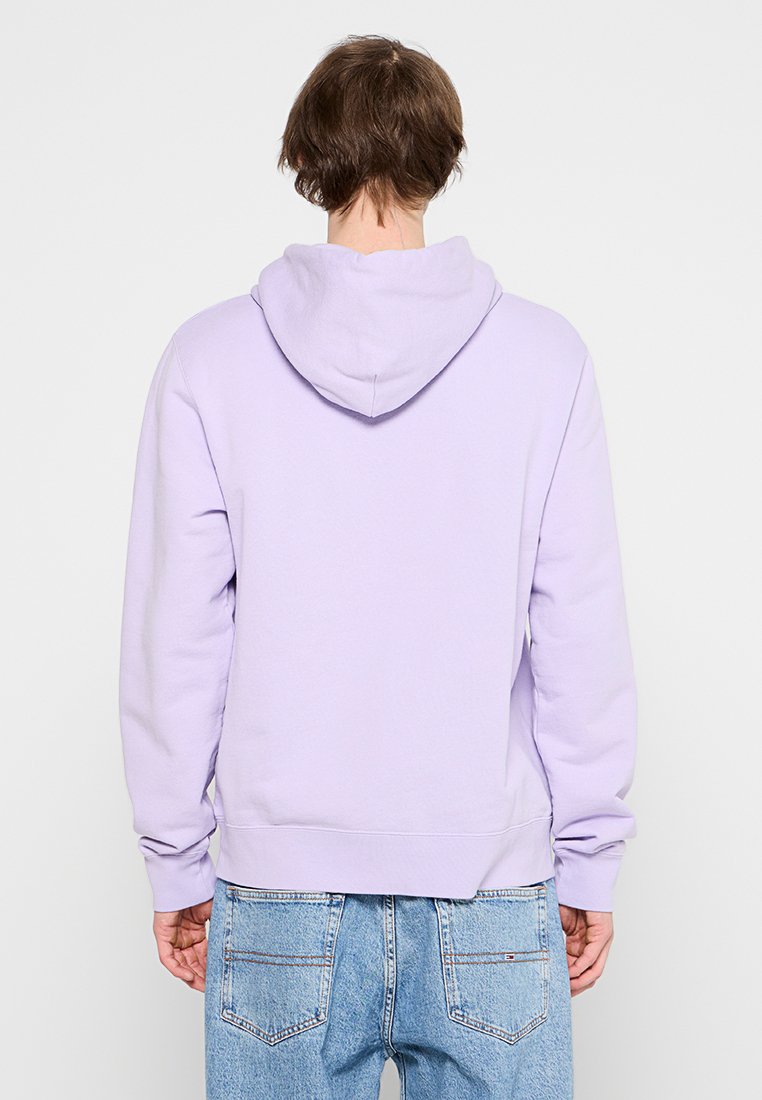 Person mit kurzen braunen Haaren, die einen helllila Hoodie und blaue Jeans tragen, von hinten vor einem schlichten Hintergrund gesehen.
