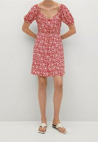 Rotes Blumenkleid mit Puffärmeln und quadratischem Ausschnitt. Der Stoff hat kleine weiße Blumen auf rotem Hintergrund. Getragen mit weißen Sandalen.