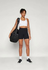 Fekete sportnadrág és fehér crop top viselése egy személy által, aki egy fekete tornazsákot tart. Az öltözet fekete sportcipővel és fehér zoknival van párosítva.