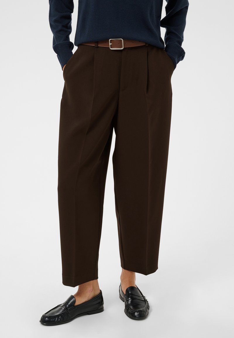 Personne portant un pantalon large marron foncé, des mocassins noirs et un haut bleu marine à manches longues, les mains dans les poches.