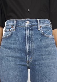 Jeans de tiro alto de color azul hechos de denim, con cinco bolsillos, herrajes de cobre y ligeras decoloraciones en los muslos para un aspecto desgastado.