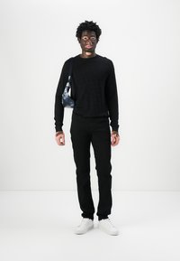 Sort striktrøje, sorte bukser, hvide sneakers, holder en lille blå taske. Bemærkelsesværdige mønstre på trøjen. Enkel, monokromt sæt.