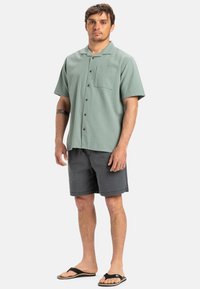 Short-sleeve, textured green shirt with a chest pocket, button closure, worn with gray shorts and black flip-flops.
