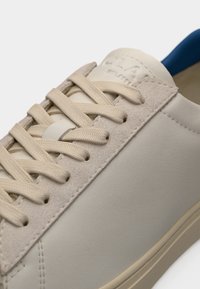 Κοντινή λήψη μπεζ αθλητικού παπουτσιού με υφασμένα κορδόνια, πάνελ από σουέτ με οπές για κορδόνια, λείες πλευρές από δέρμα και ορατή εσωτερική φόδρα σε μπλε χρώμα.