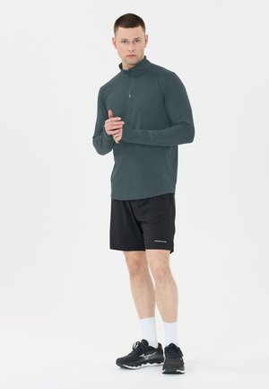 Dunkelgrünes Langarmshirt mit einem Viertel-Reißverschluss-Kragen, kombiniert mit schwarzen Shorts und schwarzen Sportschuhen. Hat eine glatte Textur und Passform.