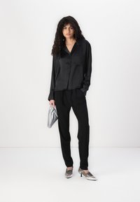 Black satin blouse with a collar, paired with black trousers. Model holds a silver, textured clutch and wears silver-heeled shoes.