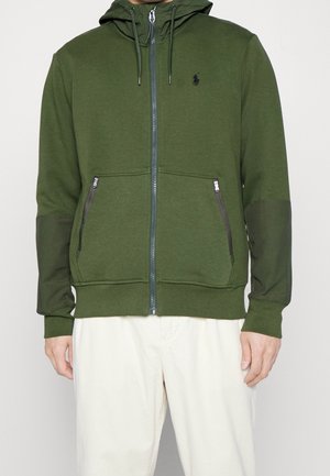 Homme portant un sweat à capuche zippé vert olive avec des poches avant zippées et un pantalon de couleur crème, sur un fond uni.