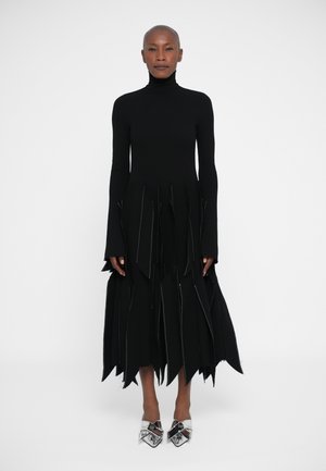 Vestido preto de mangas longas com gola alta, apresentando detalhes de franjas em camadas e costuras prateadas. Usado com sapatos de salto alto prateados e brilhantes.