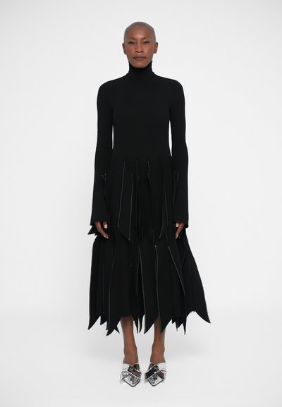 Schwarzes langärmliges Kleid mit hohem Kragen, ausgestattet mit geschichteten Fransen und silbernen Nähten. Getragen mit glänzenden silbernen High Heels.