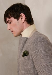 Muž v béžovom károvanom saku s vysokým smotanovým golierom a zelenou vreckovkou, pozerajúci sa smerom nadol na jednoliatom béžovom pozadí.