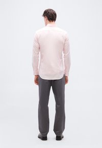 Άνδρας που στέκεται με την πλάτη, φοράει ανοιχτό ροζ μακρυμάνικο πουκάμισο, γκρι παντελόνι και μαύρα παπούτσια, σε απλό λευκό φόντο.