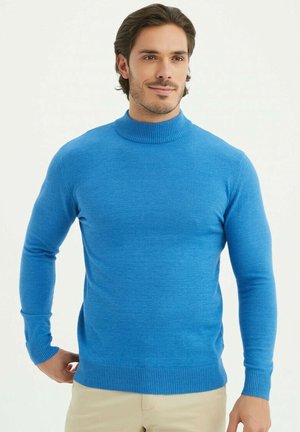 Pull-over en bleu col roulé avec une texture côtelée et des poignets, design ajusté, fabriqué dans un matériau doux, porté avec un pantalon beige, mettant en valeur une finition lisse.