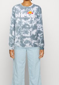 Sudadera de tie-dye azul claro con el logo blanco de "ellesse". Mangas largas, puños y bajo acanalados, textura suave. Combinada con jeans de pierna ancha azul claro.