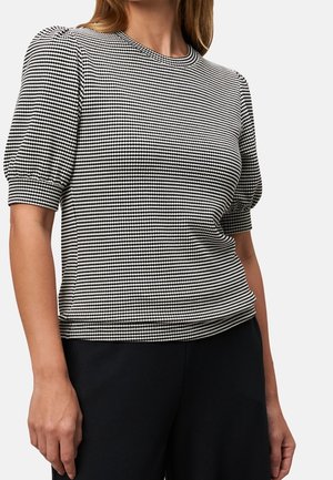Vrouw die een zwart-wit gestippeld kort mouwtopje met pofmouwen en een zwarte broek draagt, afgebeeld van nek tot heupen.