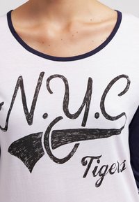 Valkoinen T-paita, jossa tummansinisiä hihoja ja rohkeat mustat tekstit "N.Y.C Tigers" sekä graafinen kuvio, joka muistuttaa tyyliteltyä asetta tai työkalua.